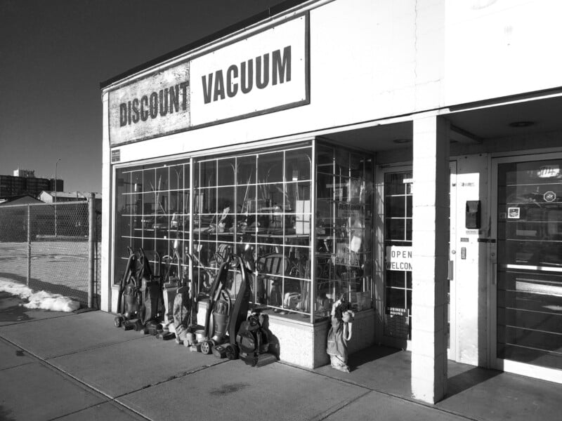 Black and white photo of a storefront labeled "Discount Vacuum," with upright vacuum cleaners lined up outside and more visible through the large window. A sign on the door says "Open." The sidewalk and part of the street are visible.