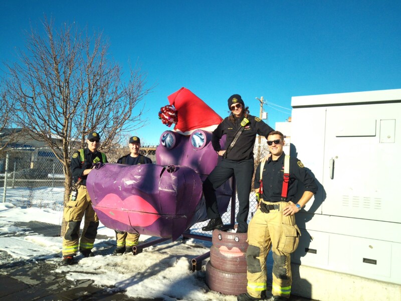 Four firefighters in uniform stand and pose beside a large whimsical purple hippo sculpture wearing a red Santa hat, on a snowy sidewalk under a clear blue sky.