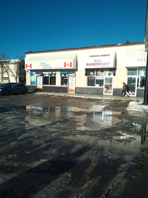 A strip mall with shops including "The Next Level" and a barbershop. Snow and ice patches cover the ground, with puddles reflecting the buildings. A person walks past the storefronts under a clear blue sky.