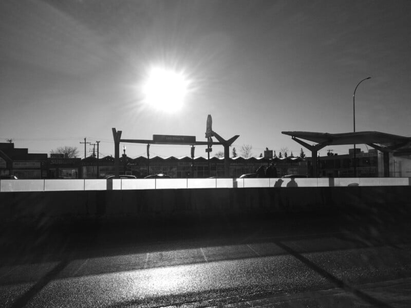 A black and white photo of a transit station at sunset with the sun shining brightly above the platform and casting long shadows. Silhouettes of shelter structures, cars, and buildings are visible in the background.