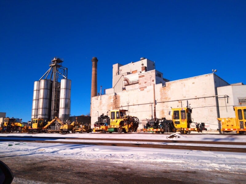 A row of yellow maintenance vehicles is parked beside train tracks in front of a large industrial building with silos and a tall smokestack on a clear, sunny winter day. Snow covers the ground.