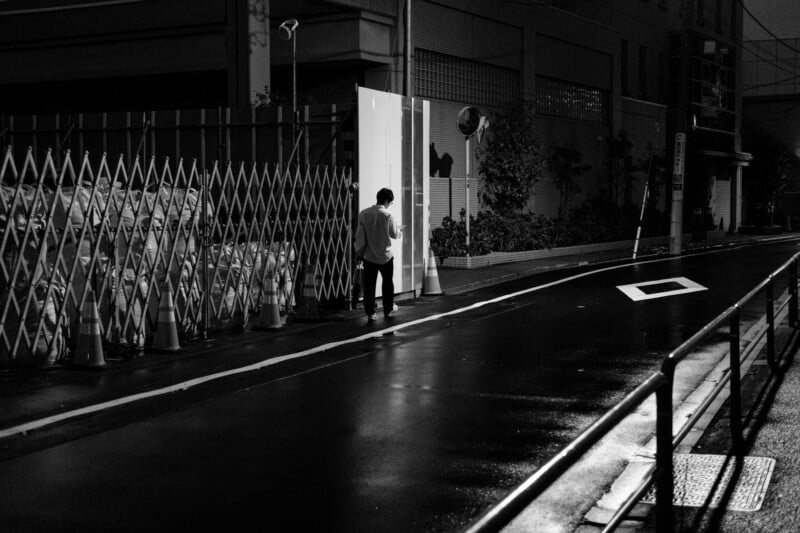 A person stands alone at night on a wet, empty street beside a tall fence and illuminated doorway. The scene is in black and white, with reflective pavement and urban surroundings.