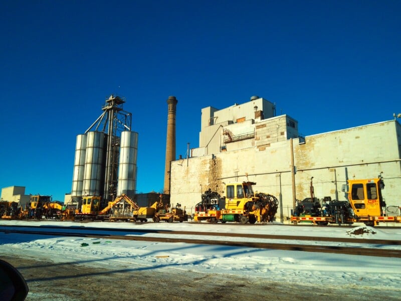 A row of yellow construction vehicles is parked in front of an industrial building with large silos and a tall chimney on a clear, sunny winter day. Snow covers the ground and train tracks run across the scene.