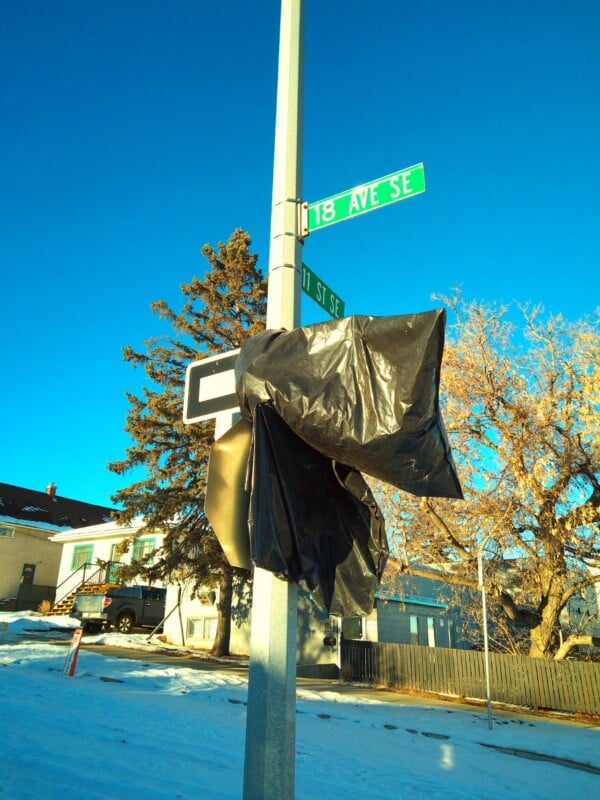 A street sign at the corner of 18 Ave SE and 11 St SE is covered with a black plastic bag. Snow is on the ground, and houses and trees are visible in the background under a clear blue sky.