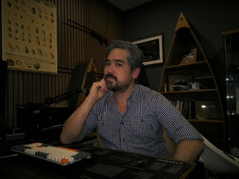 A man with gray hair and a beard sits at a desk with a keyboard, resting his chin on his hand. Behind him are shelves with books, a framed photo, a wooden triangular structure, and a poster of arrowheads.