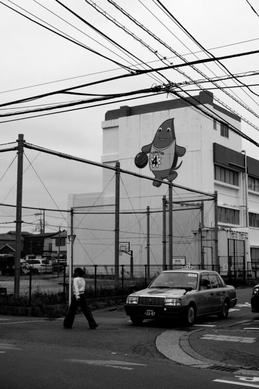 A black-and-white street scene shows a person crossing near a taxi, with power lines overhead. A large cartoon fish mascot holding a basketball is painted on the building in the background.