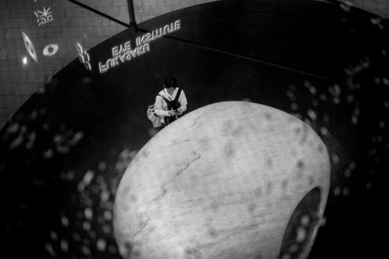 A person in dark clothing sits alone on a large, smooth, white stone in a dimly lit space, surrounded by abstract light reflections and shadowy patterns.