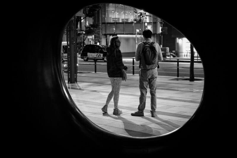 Two people stand and talk on a city sidewalk at night, seen through a large circular framing. Both cast long shadows on the pavement, with streetlights and a parked car in the background. The photo is in black and white.