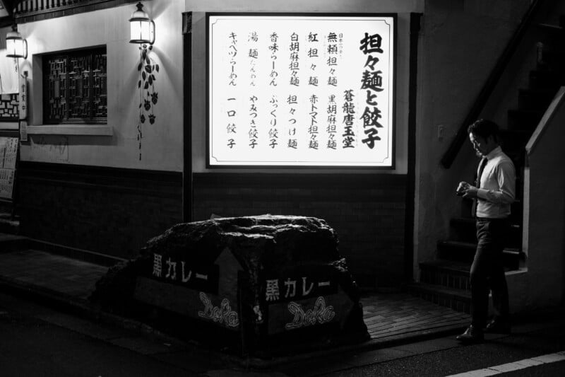 A man stands outside at night near a lit restaurant sign with Japanese text. The area is dimly lit, with lanterns on the building and a large rock in front labeled “黒カレー” (black curry).