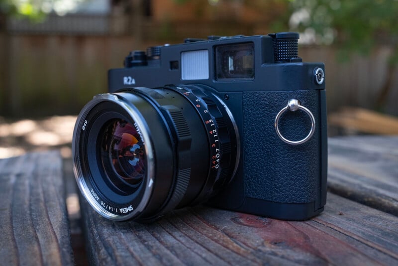 A black rangefinder camera with a large lens rests on a weathered wooden table outdoors, with a blurred background of a fence and greenery.