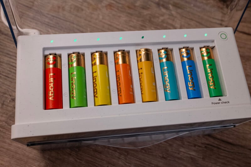 Eight rechargeable batteries in various colors (red, green, orange, yellow, and blue) are inserted in a white charging station. Green indicator lights above each battery show charging status. The charger rests on a wooden surface.