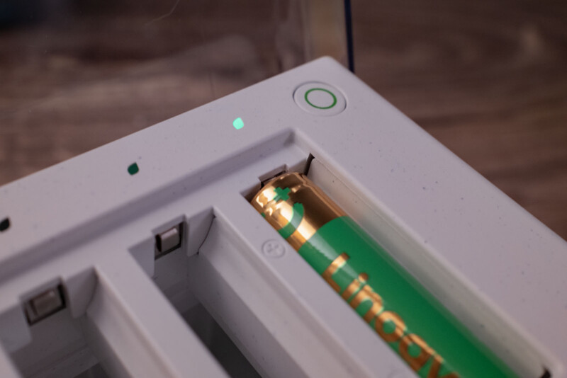 A close-up of a battery charger with a green and gold AA battery inserted. A small green indicator light is illuminated next to a circular power button on the white charger.