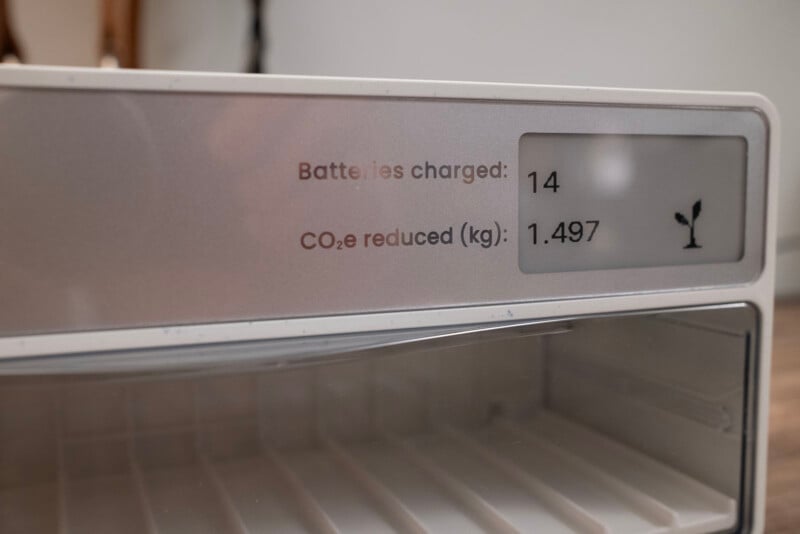 Close-up of a display panel showing “Batteries charged: 14” and “CO₂e reduced (kg): 1.497” with a small plant icon, mounted above an empty shelf or compartment.