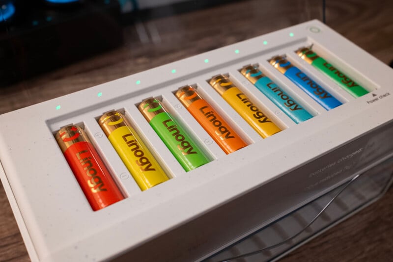 A white battery charger with eight colorful Linogy batteries—orange, green, yellow, and blue—lined up inside it. Small green LED lights above each slot indicate charging status. The charger sits on a wooden surface.