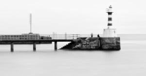 Black and white photo of a pier with stairs leading into the water, a striped lighthouse on the right, another light structure in the distance, and blurry figures standing on the pier. The water appears smooth and misty.