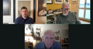Screenshot of a video call with three men. The top left man wears a dark shirt in front of framed photos; the top right man has white hair, glasses, and a beard, with bookshelves behind him; the bottom man wears glasses in a cluttered room.