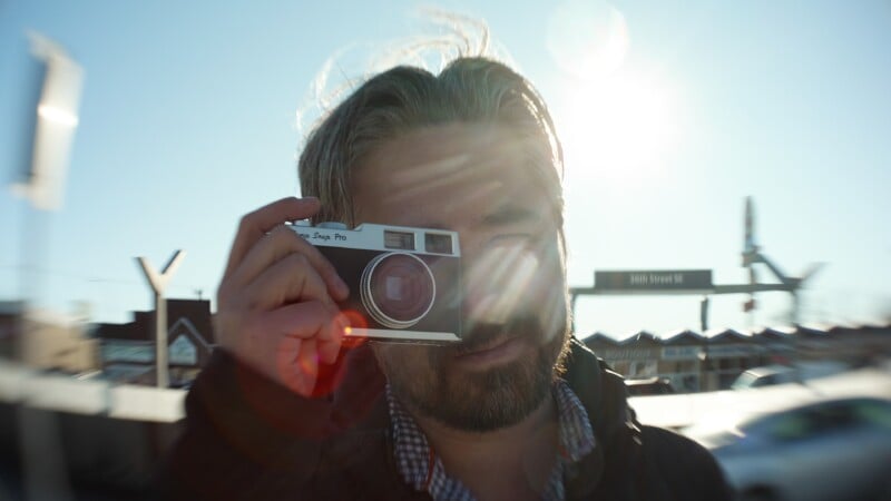 A man with light brown hair and a beard holds a vintage camera up to his face outdoors, with sunlight and lens flare partially obscuring his face. A street sign and buildings are visible in the blurred background.