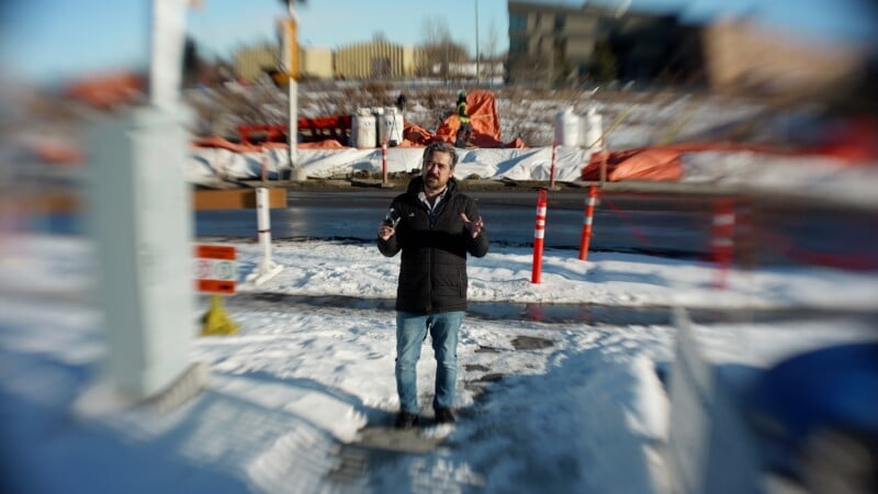 A man in a dark jacket and jeans stands on a snowy sidewalk near a street with construction materials, orange barriers, and equipment in the background. The image has a blurred, vignette effect around the edges.