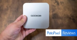 Hand holding a compact silver device with the brand name "GEEKOM" on top. The background is a wooden surface, and the "PetaPixel Reviews" logo is visible at the bottom right.
