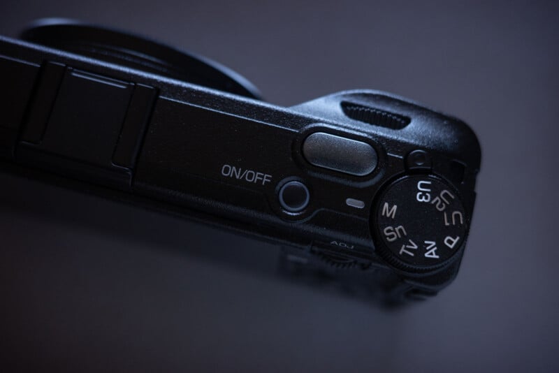Close-up of a black digital camera's top view, showing the on/off button, a control dial with various mode settings, and part of the lens against a dark background.