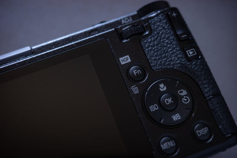 Close-up of the back of a digital camera showing buttons, dials, and part of the LCD screen, with textured grip and mode controls visible.