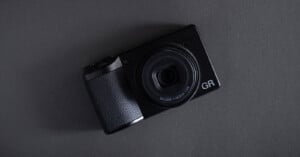 A black compact digital camera with a large lens, labeled "GR," placed on a dark gray surface at a slight angle.