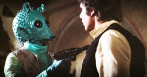 A green alien with textured skin and large black eyes points a blaster at a man with brown hair, wearing a white shirt and black vest. The scene has a tense atmosphere, set indoors with soft lighting.