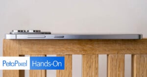A side view of a silver smartphone with prominent camera bumps, resting on a wooden surface. The image features the "PetaPixel Hands-On" logo in the bottom left corner.