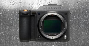 A Hasselblad camera body without a lens is shown against a wet, rain-covered glass background. Water droplets cover the surface behind the camera.