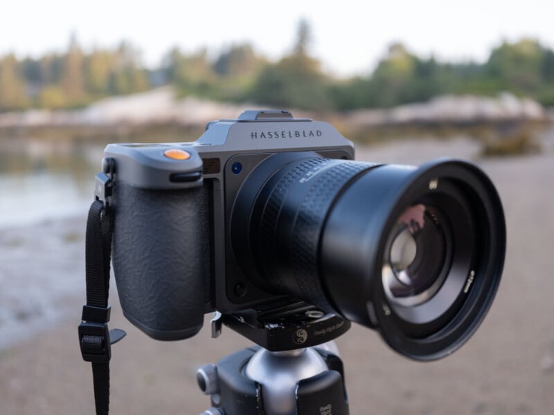 A Hasselblad camera mounted on a tripod is set up outdoors, with a blurred background of trees, water, and rocks.