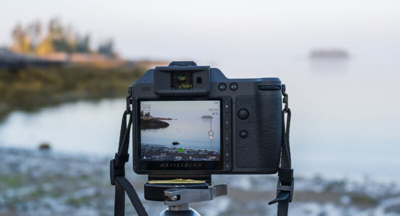 A digital camera on a tripod points at a calm lakeside scene, displaying the landscape on its screen. The background is blurred, showing water, shoreline, and distant trees under a soft, bright sky.