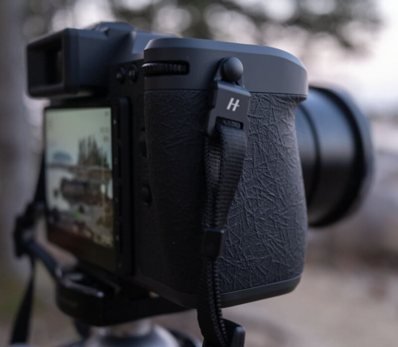 Close-up of a black digital camera mounted on a tripod, showing a textured grip and a screen displaying an outdoor scene with trees and water in the background.