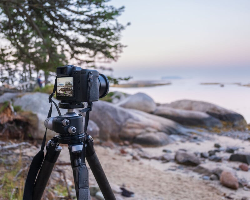 A camera on a tripod is set up on a rocky shoreline, facing a calm, misty body of water with distant rocks and trees. The camera’s display shows the landscape it is capturing.