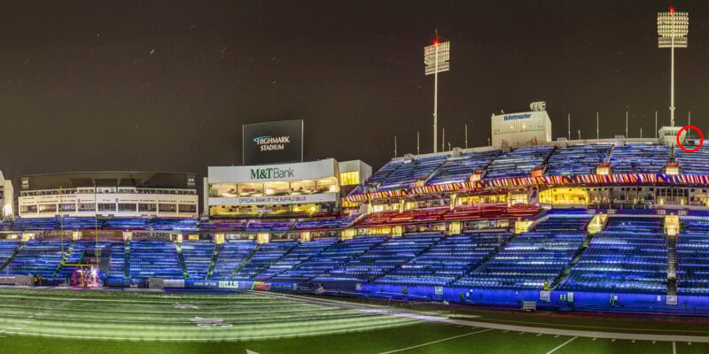 A wide-angle night view of a brightly lit empty football stadium with blue seats, spotlights, and large video boards, including a sign for M&T Bank. The field is partially visible in the foreground.