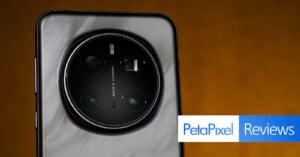 Close-up of a smartphone's rear camera setup, featuring a circular module with multiple lenses. The background is a warm brown tone. The Petapixel logo and the word "Reviews" appear at the bottom right.