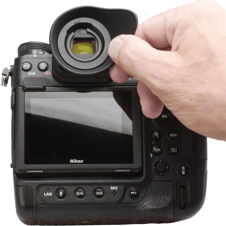 A hand is removing a rubber eyecup from the viewfinder of a Nikon digital camera, which is shown from the back with its screen and various buttons visible.