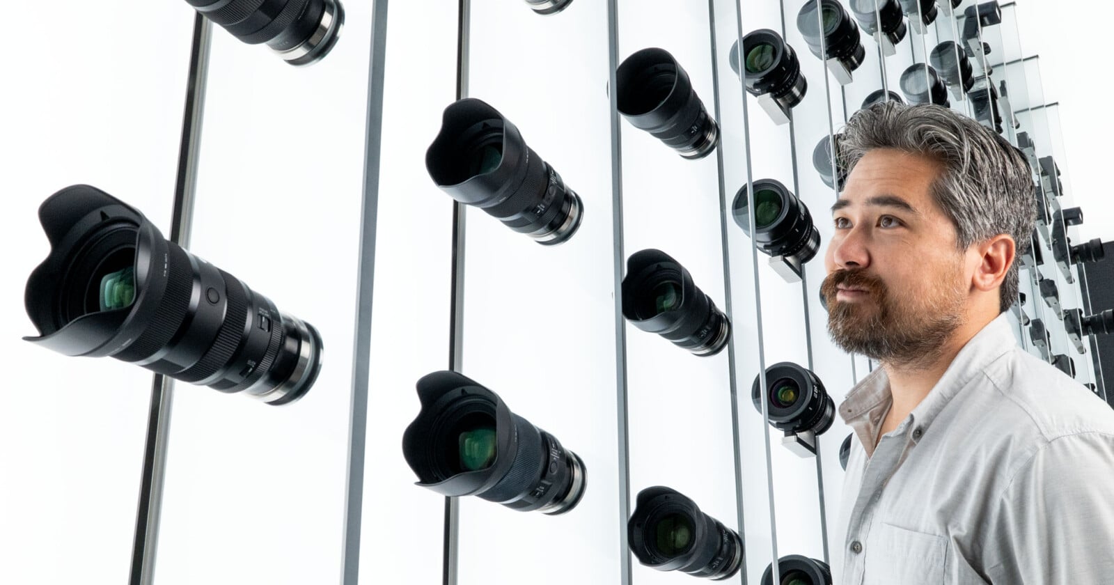 A man with gray hair and a beard stands in front of a wall lined with rows of camera lenses, some of which are reflected in the mirrored surface behind him.