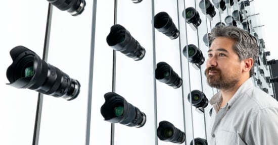 A man with gray hair and a beard stands in front of a wall lined with rows of camera lenses, some of which are reflected in the mirrored surface behind him.