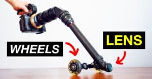 A camera mounted on a rig with skateboard wheels and a lens at the end, labeled "WHEELS" and "LENS" with arrows pointing to each part. The setup rests on a wooden surface against a plain background.