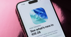 A smartphone screen displays an app update notification for iOS 26, featuring a blue and green abstract icon with the number 26 and the headline “See what’s new in iOS 26.” The background is pink and shiny.