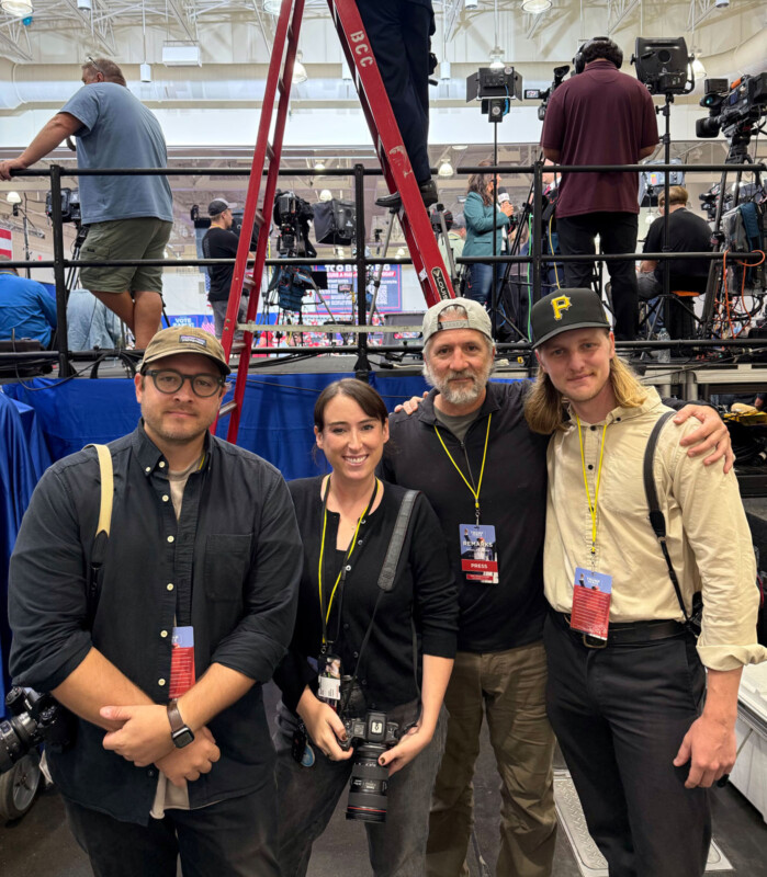 Four people with cameras and press badges stand and smile for a photo in front of a press area with equipment, ladders, and other media personnel in the background. The setting appears to be an indoor event or convention.