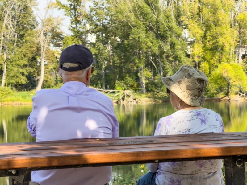 An elderly man and woman sit on a wooden bench by a calm lake, facing away from the camera. They are surrounded by trees and greenery on a sunny day.