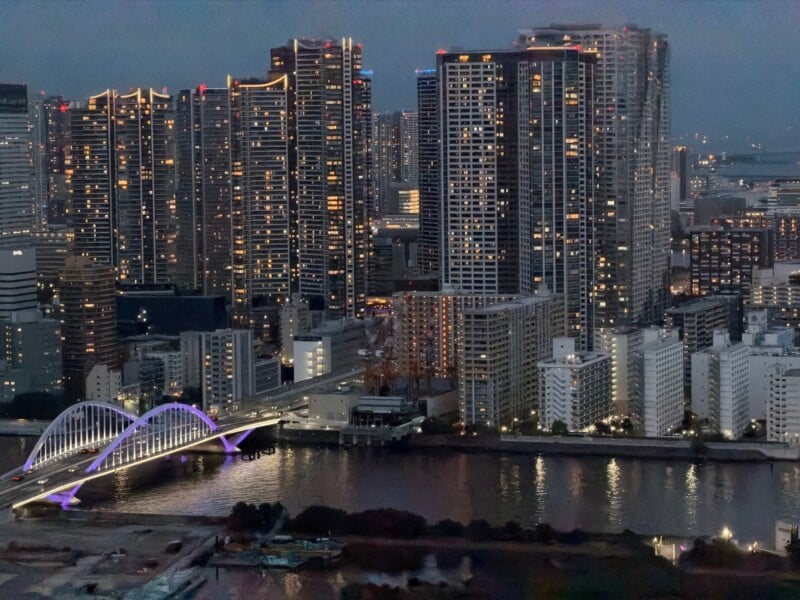 A cityscape at dusk showing tall illuminated skyscrapers, a lit-up arched bridge over a river, and reflections of city lights on the water. The sky is dim, indicating early evening.