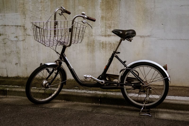 A black bicycle with a large metal basket attached to the front is parked on a street next to a worn, beige concrete wall. The bike is slightly angled, resting on its kickstand.