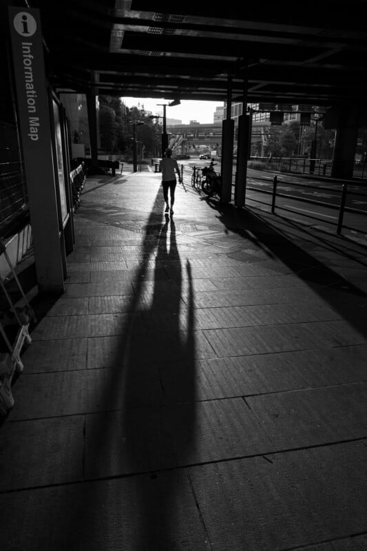 A person walks away from the camera on a sidewalk, casting a long shadow in the low sunlight under an overpass near an "Information Map" sign; bicycles are parked ahead.