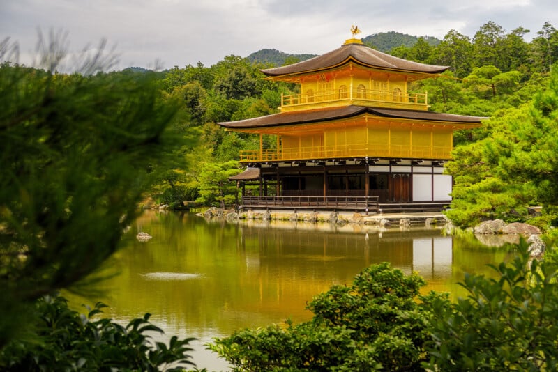 A golden pavilion sits beside a calm pond surrounded by lush green trees and bushes, with mountains visible in the background under a cloudy sky.