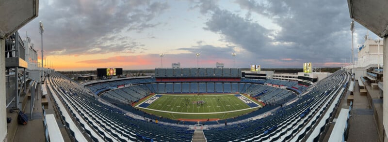 Wide-angle view of an empty football stadium at sunset, with rows of blue seats surrounding a green field marked "BILLS" in the end zones. The sky is partly cloudy, and stadium lights are illuminated.