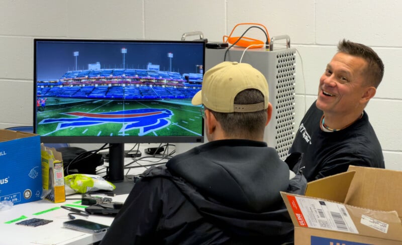 Two men sit at a desk with computer equipment. One faces a large monitor displaying a football stadium field with the Buffalo Bills logo. The other man is smiling and looking at the camera. Boxes and snacks are on the desk.
