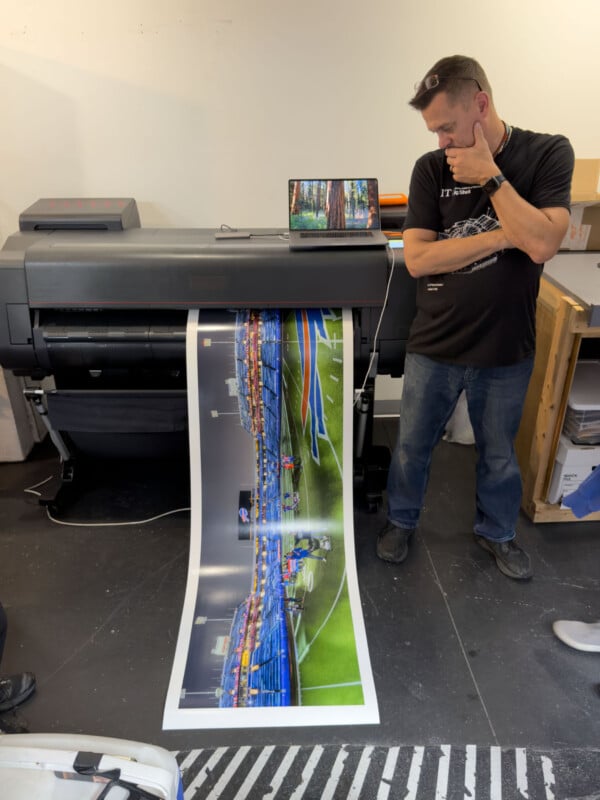 A man stands thoughtfully beside a large-format printer producing a colorful panoramic print of a stadium or sports field. A laptop is placed on top of the printer, and boxes are stacked nearby in the workspace.