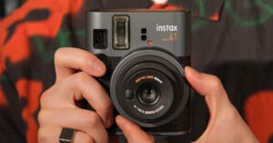 A person holds a black Instax Mini 40 instant camera, showing the lens and flash. The camera has a textured body, and the person is wearing a patterned shirt and a ring on their right hand.